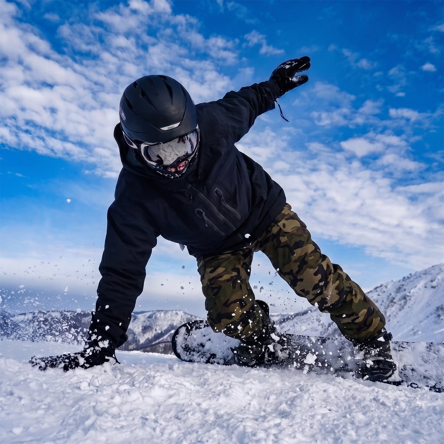 Lifestyle action photo of a rider carving on the XCMAN Men's Ember Nocturne All Mountain Snowboard down a snowy mountain slope under a partly cloudy sky