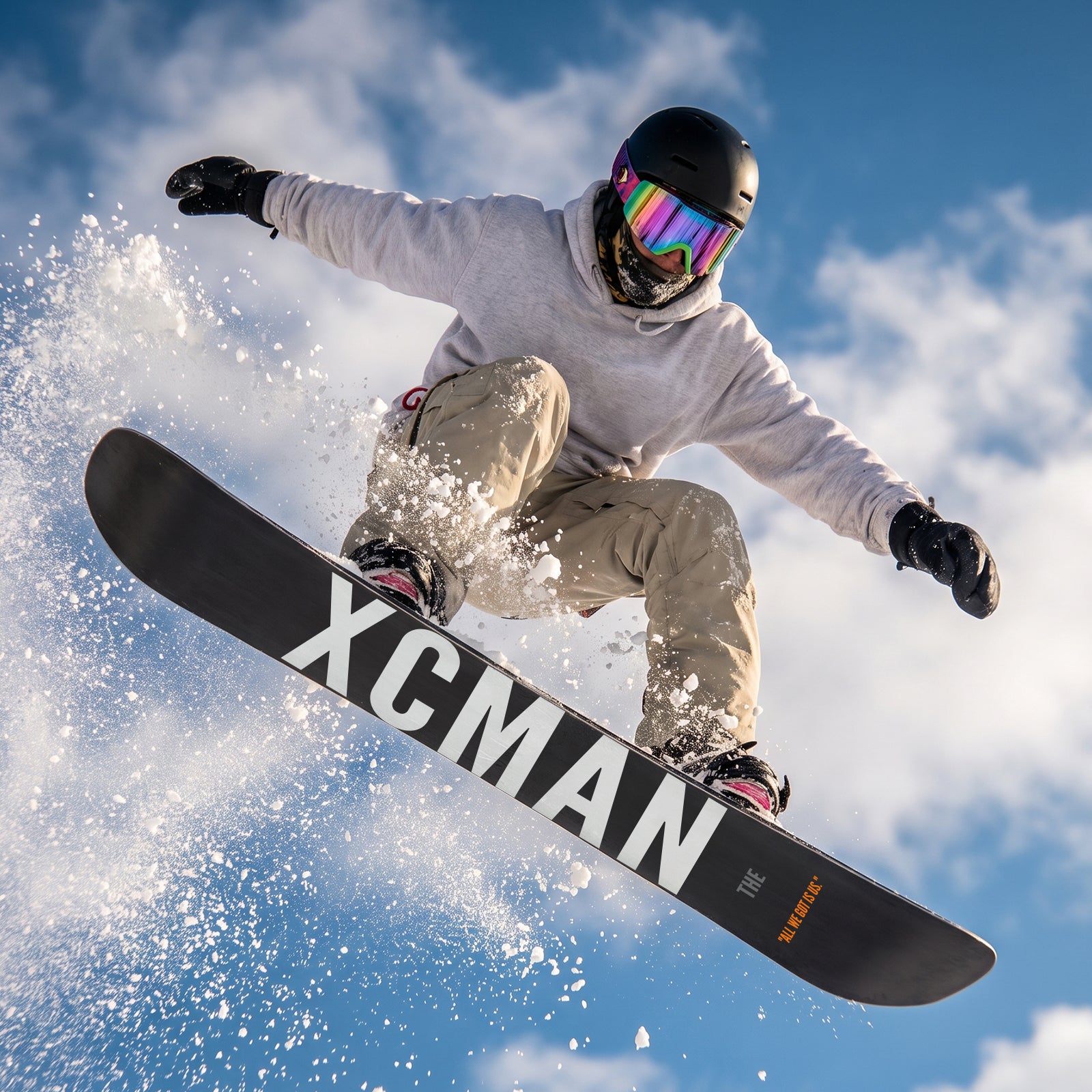 Lifestyle action photo of a snowboarder jumping on the XCMAN Men's Cloudy Forest All Mountain Snowboard against a snowy mountain and blue sky backdrop